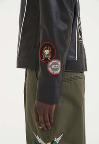 Giubbotto di pelle nera con toppe ricamate rosse e bianche sulle maniche, dal design aderente e con zip contrastanti.