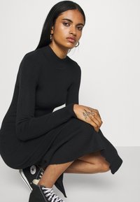 Robe en tricot côtelé noire avec un col haut et des manches longues, présentant un ourlet évasé et assortie à des baskets montantes noires.
