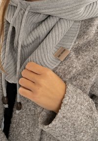 De hand van een vrouw die een lichtgrijze gebreide sjaal met houten kralen over een beige texturen jas aanpast.