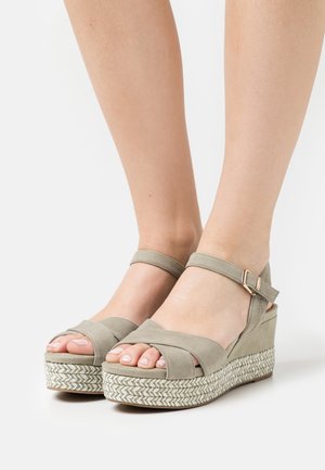 Jasnoszare sandały na koturnie o miękkiej zamszowej teksturze, z krzyżującymi się paskami, zapięciem na kostce oraz plecioną podeszwą zygzakowatą.
