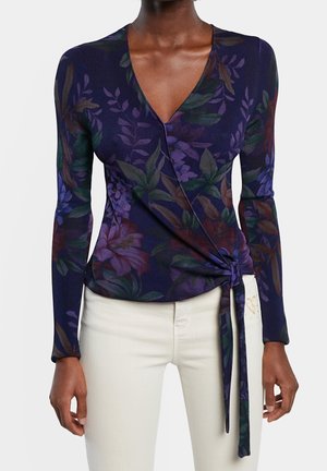 Vrouw die een donker bloemleer wikkelblouse met lange mouwen en zijstrik draagt, gecombineerd met lichtgekleurde broek.