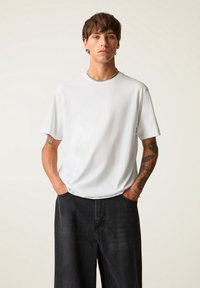 White short-sleeved t-shirt with a crew neck, made of soft fabric. Paired with dark, loose-fitting pants, featuring a minimalist design.