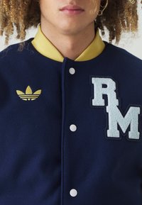 Mornarska volnena jakna z rumenim ovratnikom, srebrnimi gumbi s kavljem, vezenim "RM" v svetlo modri barvi in logotipom Adidas v zlati barvi na levi strani.
