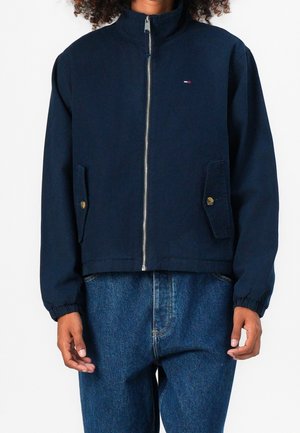 Person trägt eine marineblaue Jacke mit Reißverschluss, Knopfleistentaschen und elastischen Bündchen, kombiniert mit blauen Jeans.