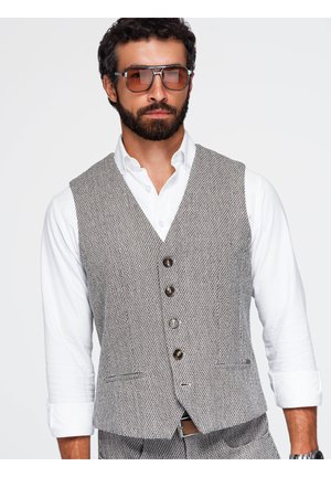 Man met baard en zonnebril draagt wit overhemd en grijs gemêleerd vest met knopen tegen een effen achtergrond.