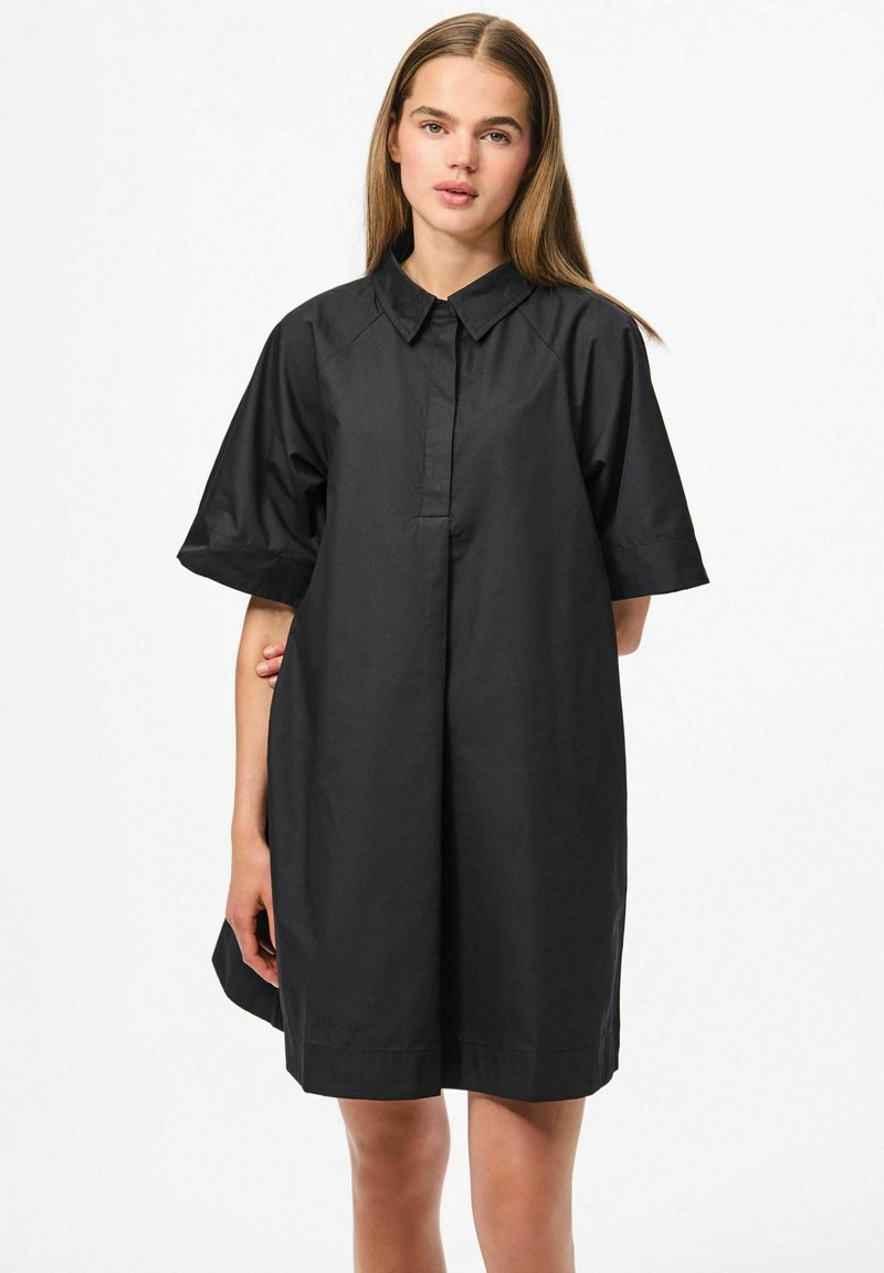 Abito camicia nero oversize realizzato in tessuto morbido, dotato di colletto a punta, maniche corte e orlo dritto senza chiusure visibili.
