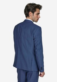 Junger Mann in einem maßgeschneiderten blauen Anzug, von hinten betrachtet, mit nach hinten gekämmten Haaren und einer selbstbewussten Haltung, vor einem weißen Hintergrund.