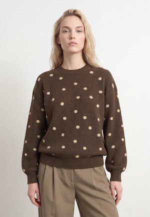 Femme aux longs cheveux blonds portant un pull marron à pois beige et un pantalon plissé beige, debout sur un fond blanc uni.
