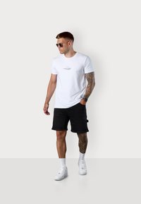 Weißes Baumwoll-T-Shirt mit bedrucktem Text, schwarze Denim-Shorts, weiße Sneaker und Sonnenbrille; männliche Figur mit Armtattoos.
