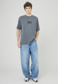 T-shirt en coton gris avec un imprimé noir "NIRVANA", associé à un jean baggy bleu clair. Le modèle porte des baskets grises et noires.