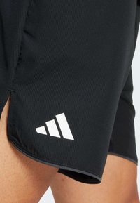Detail černých sportovních šortek s bílým logem Adidas na stehně, které nosí osoba s viditelnou pletí a chlupy na nohou.