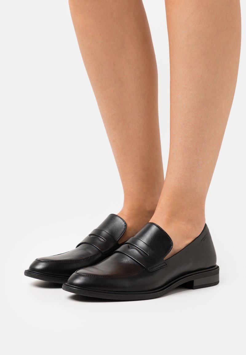 Mocassins penny en cuir noir portés pieds nus, avec un talon bas et une finition lisse et polie.