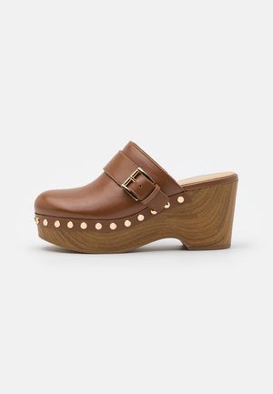 Zueco de cuero marrón con una correa ancha y hebilla dorada, apoyado sobre una plataforma de madera curvada adornada con pequeños tachuelas redondas.