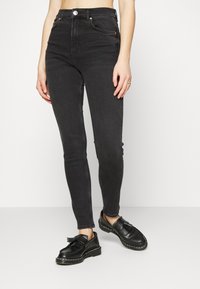 Jeans de ganga pretos de cintura alta com um corte justo, apresentando um fecho na parte da frente, dois bolsos frontais e um desbotado subtil. Combinados com sapatos pretos.