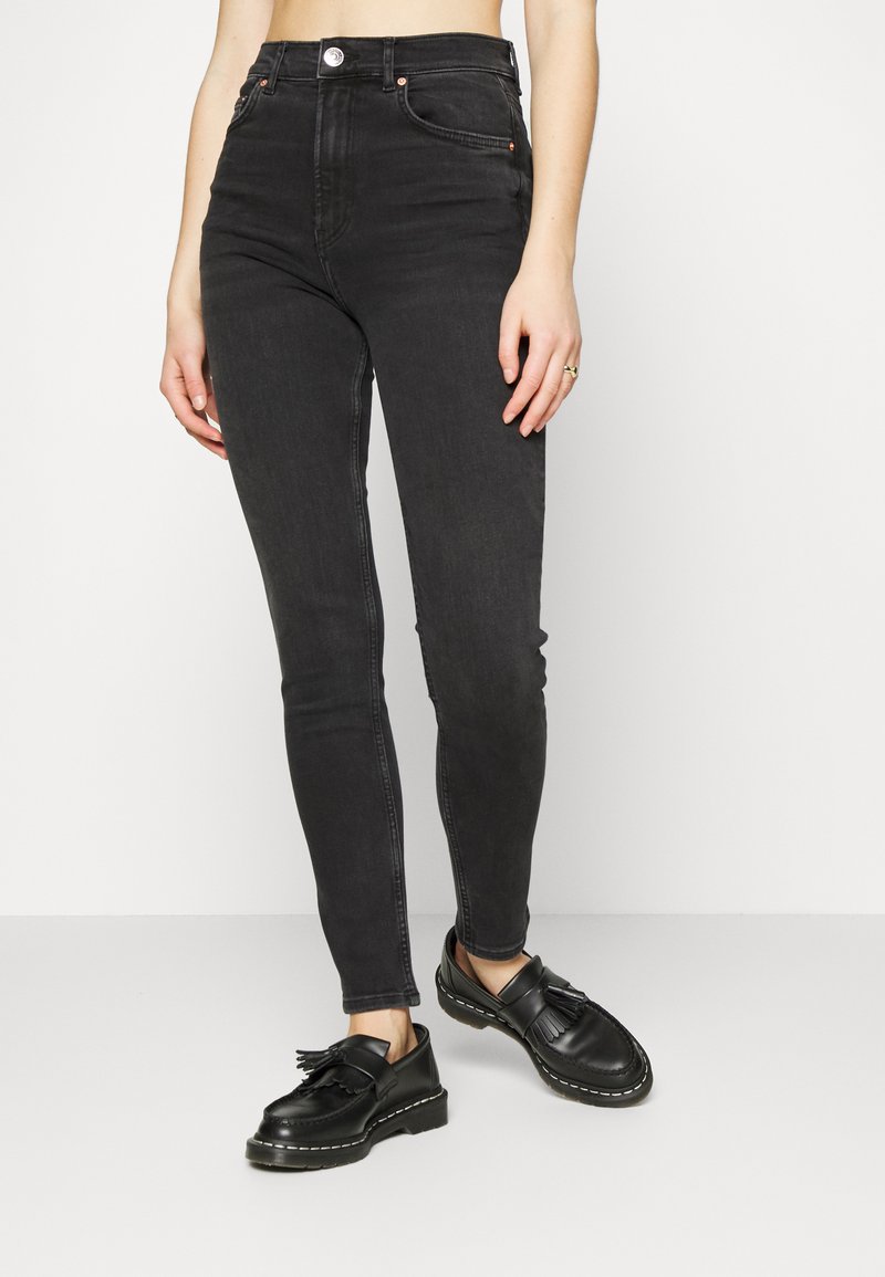 Jeans de ganga pretos de cintura alta com um corte justo, apresentando um fecho na parte da frente, dois bolsos frontais e um desbotado subtil. Combinados com sapatos pretos.