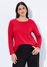 Rotes langärmliges T-Shirt aus Baumwolle. Mit Rundhalsausschnitt und lässiger Passform, kombiniert mit schwarzen Hosen. Einfaches Design, keine Muster oder Akzente.