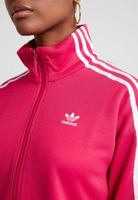 Różowa kurtka dresowa z zamkiem błyskawicznym, z białymi paskami na ramionach i rękawach. Posiada haftowane białe logo Adidas na piersi.