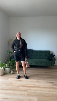 T-shirt preto de mangas compridas com detalhes em renda, calções pretos de cintura alta e sapatos pretos. Segurando uma bolsa bege, com um sofá verde ao fundo.