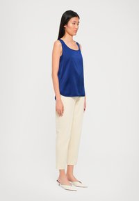 Woman stands wearing sleeveless blue satin top, cream straight-leg trousers, and pointed cream low-heeled shoes against plain background.