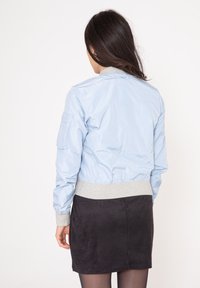 Ljusblå bomberjacka med ribbade grå ärmar och fåll. Har sidofickor och en slät, glänsande yta. Bärs tillsammans med en svart mockakjol.