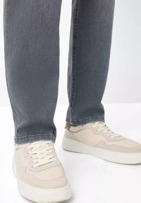 Beige sneakers with textured suede and smooth leather panels, white laces, and a thick rubber sole, paired with gray denim jeans.