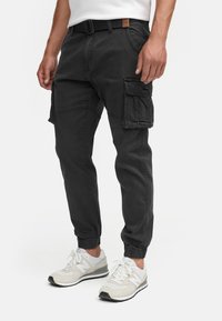 Homme portant un pantalon cargo noir avec de multiples poches et des ourlets élastiqués, associé à des baskets blanches et beiges.