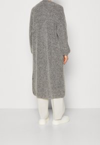 Personne debout, portant un long manteau gris texturé, un pantalon blanc à jambes larges et des baskets blanches, sur un fond clair uni.