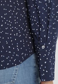Chemise bleu marine avec un petit motif géométrique en bleu clair et blanc. Elle possède des manches longues avec des poignets à boutons et un ourlet incurvé.