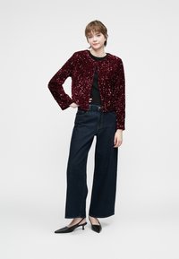 Burgundijas krāsas sekvīnu žakete ar apaļu kakla izgriezumu, uzvilkta virs melnas blūzes. Kombinēta ar platām, tumšām džinsu biksēm un melniem siltajiem papēžiem.