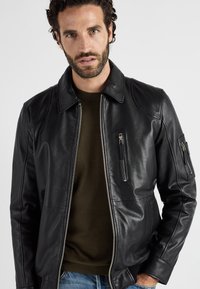 Schwarze Lederjacke mit Kragen, ausgestattet mit zwei Reißverschlusstaschen auf der Brust und gerippten Bündchen, getragen über einem olivgrünen Pullover.