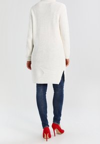 Langer weißer Strick-Cardigan mit einem strukturierten Muster, seitlichen Schlitzen und einem ungleichmäßigen Saum, kombiniert mit blauen Jeans und roten hohen Schuhen.
