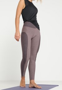 Kvinde der står barfodet på en lilla yogamåtte iført to-farvede lilla leggings og en ærmeløs sort top.