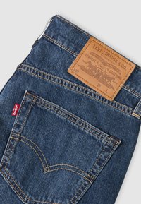 Poche arrière d'un jean en denim bleu avec une étiquette en cuir marron de Levi Strauss & Co. et une étiquette en tissu rouge Levi’s sur la couture latérale.
