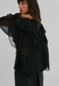 Černá průsvitná blůza s vrstvenými volánky, dlouhými rukávy a volným střihem. V kombinaci s pruhovanými kalhotami, které zdůrazňují texturu a pohyb.