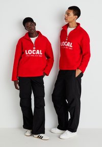 Δύο άτομα φορούν κόκκινα φλις πουλόβερ με το κείμενο «LOCAL CPH STARS, EST. 1984», μαύρο παντελόνι και λευκά αθλητικά παπούτσια, στέκονται μπροστά σε έναν λευκό τοίχο.