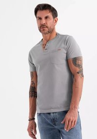 Camisa henley de hombre en gris, de manga corta y fabricada en algodón, con un escote abotonado y un bolsillo en el pecho. Corte casual con detalles de tatuajes visibles.