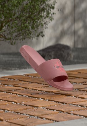 Sandalo slide rosa Calvin Klein sospeso sopra una superficie esterna in legno e ciottoli con vegetazione e sfondo in cemento.