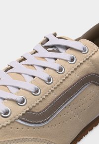 Baskets en daim beige avec lacets blancs, œillets argentés et une bande ondulée marron sur le côté.