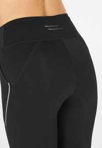 Leggings sportivi neri con vita alta, caratterizzati da cuciture flatlock, una superficie liscia e dettagli riflettenti sui lati.