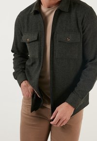 Camisa chaqueta de mezcla de lana verde oscuro con dos bolsillos en el pecho, botones de hardware de bronce y cierre de cremallera, llevada sobre una camiseta de cuello redondo beige con textura.