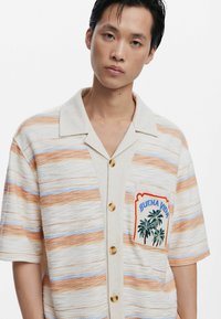 Korte mouwen shirt in crème met oranje en blauwe strepen, voorzien van een borstzak met geborduurde palmen en de tekst "Buena Vida".