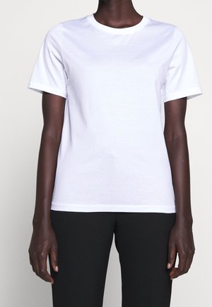 Personne portant un t-shirt blanc uni à manches courtes et un pantalon noir, debout avec les bras détendus le long du corps sur un fond neutre.