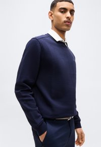 Νέος άνδρας που φοράει μπλε ναυτικό φούτερ πάνω από λευκό πουκάμισο με γιακά και μπλε ναυτικό παντελόνι, στέκεται με το ένα χέρι στην τσέπη μπροστά σε απλό φόντο.