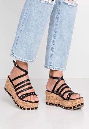 Fødder iført lyseblå revnede jeans og sorte strop-platform kilesandaler med korksåler og guldsnitter.