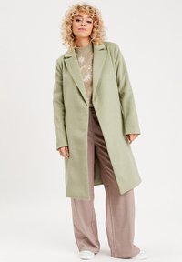 Long manteau en laine vert menthe avec un col cranté, associé à un pull à motifs, un pantalon large beige clair et des chaussures blanches.