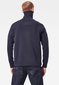 Felpa blu navy con collo alto e zip frontale, caratterizzata da un tessuto testurizzato e una vestibilità rilassata. Abbinata a jeans in denim scuro.