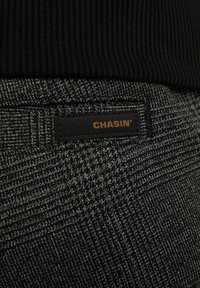 Detailní záběr šedých vzorovaných kalhot s černým štítkem s nápisem "CHASIN'" pod černým žebrovaným topem.
