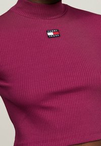 Detailní záběr na fialový žebrovaný top s vysokým límcem a malou obdélníkovou nášivkou Tommy Jeans na hrudi.