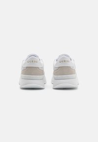 Adidași albi și gri cu accente din piele întoarsă, având o talpă intermediară texturată și un logo "GUESS" aurit pe călcâi.