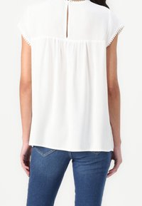 Blusa bianca con maniche corte, rifiniture testurizzate sulle maniche, dettaglio a oblò sul retro e pieghe sulla parte posteriore, abbinata a jeans blu.